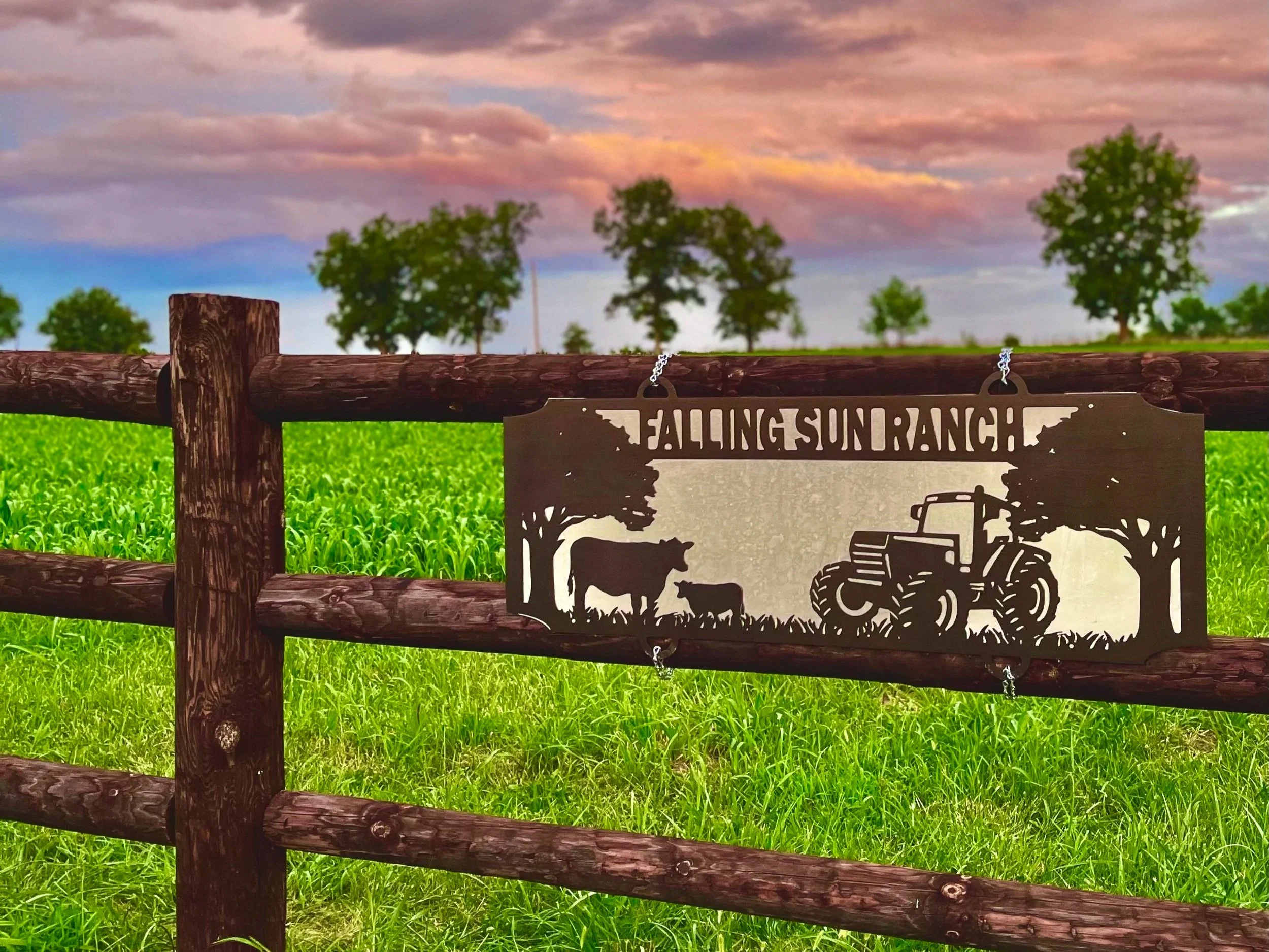 Sign for Falling Sun Ranch displayed on a wooden fence, featuring silhouettes of a tractor, cow, and trees, with a lush green field and a colorful sky in the background.