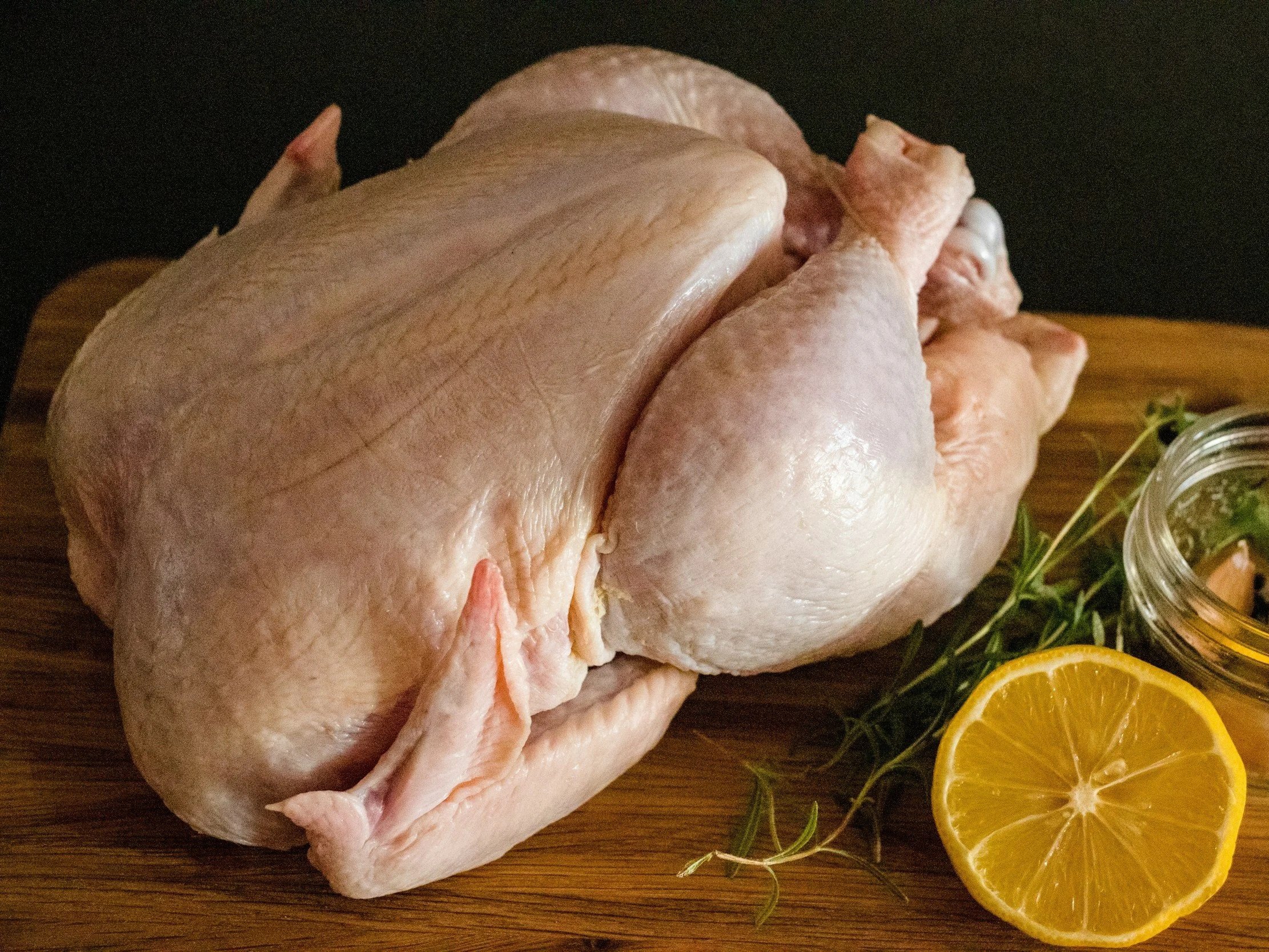 unsplash-image-9zLa37VNL38 Raw whole chicken on a wooden cutting board, accompanied by fresh herbs and a sliced lemon, ideal for cooking and meal preparation.