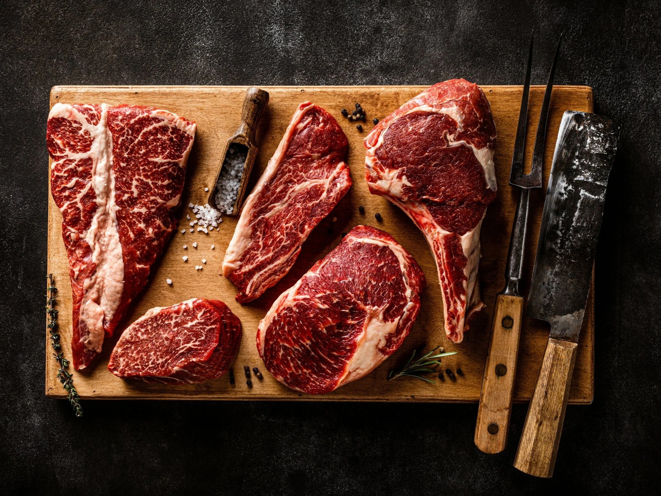unsplash-image-cEJGctX_DgI (1) Assorted cuts of raw marbled beef displayed on a wooden cutting board, featuring ribeye, sirloin, and tenderloin steaks, accompanied by a wooden scoop of salt and seasoning, with butcher tools nearby.