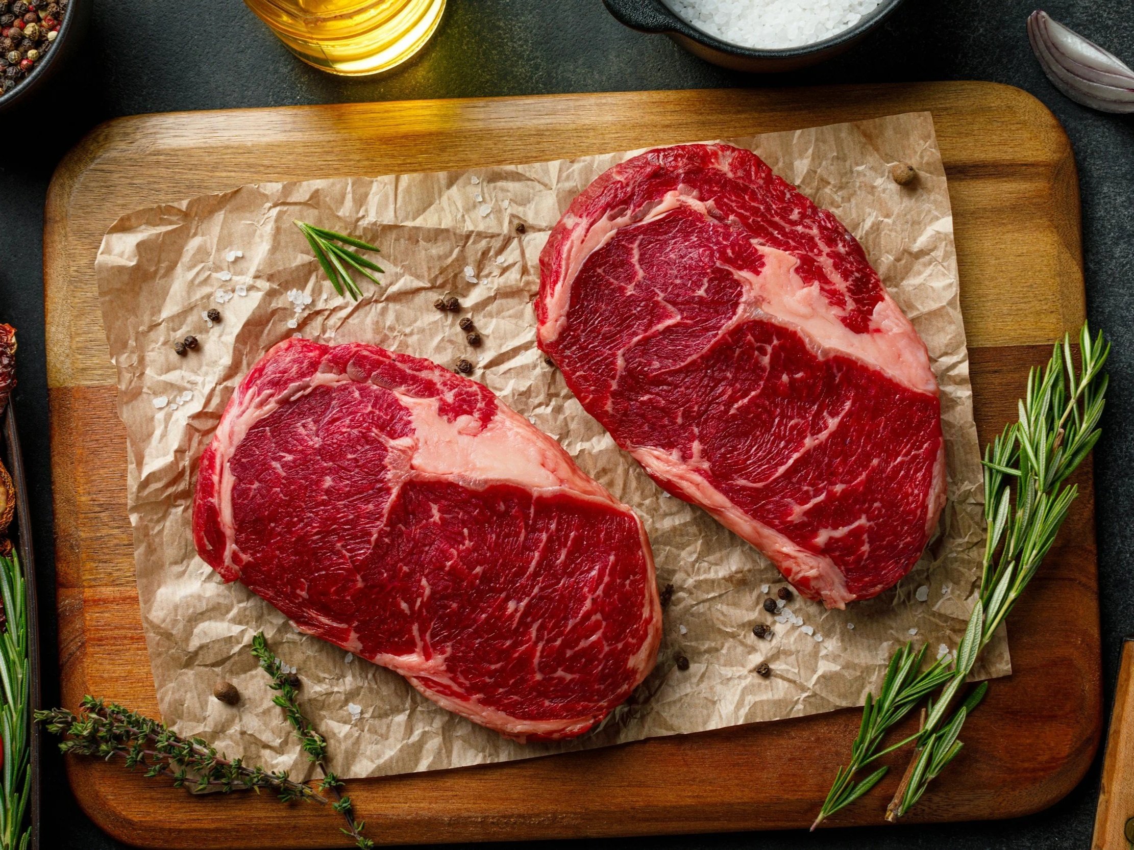 unsplash-image-x--wK7akQWw Fresh ribeye steaks on parchment paper, garnished with herbs and spices, displayed on a wooden cutting board. Ideal for grilling or roasting, these cuts showcase rich marbling and vibrant red color, perfect for meat lovers.